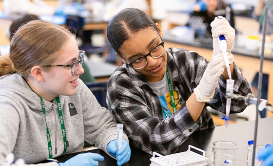 Two students participate in a science lesson. One student in a grey sweatshirt and blue rubber gloves looks on as the other student in a black and white flannel shirt and white rubber gloves pipes a material into a tube.