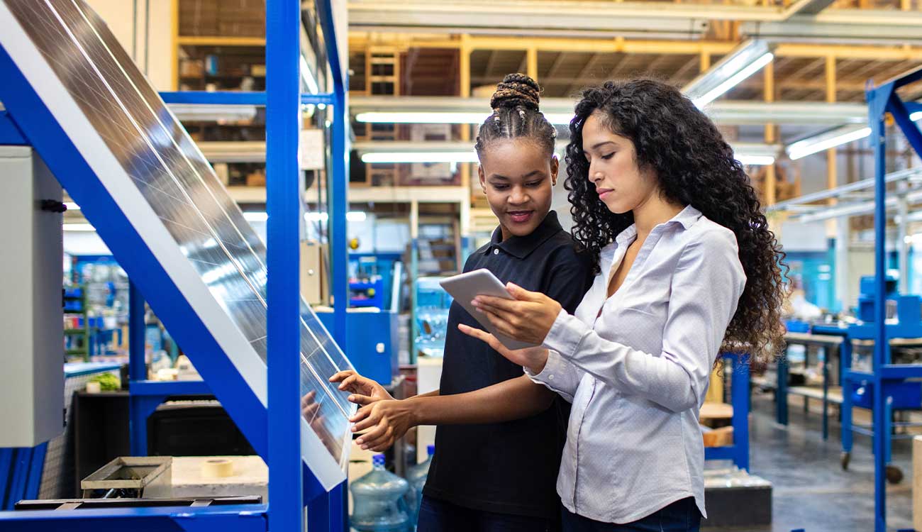 Two workers in a solar panel factory. One worker holds a tablet for the other to look. This image depicts "Connecting School and Career in Rural Communities"