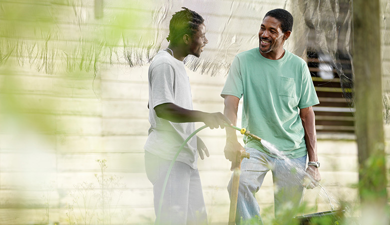An older man and a younger man talking, with the younger man holding a watering hose, in an outdoor setting.