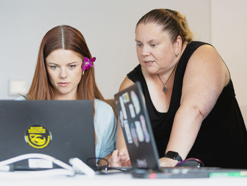 Woman leaning toward a youth at a desk with two laptops in a teaching setting.