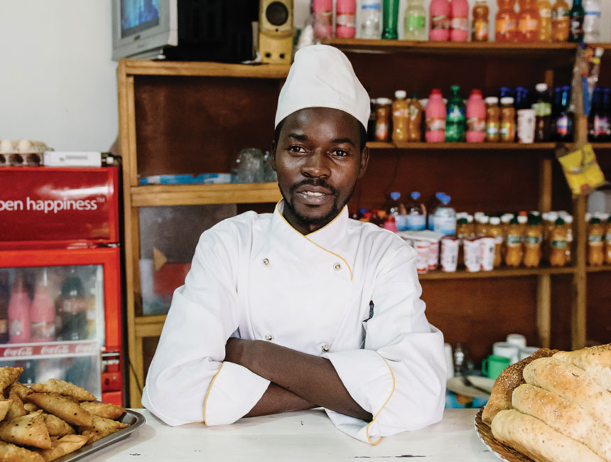 Man in chef uniform facing the camera, with kitchen ingredients behind him.