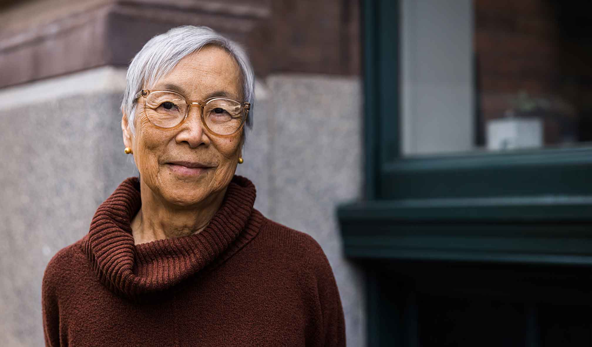 Healthy-looking older woman smiling at the camera, standing in front of a building.