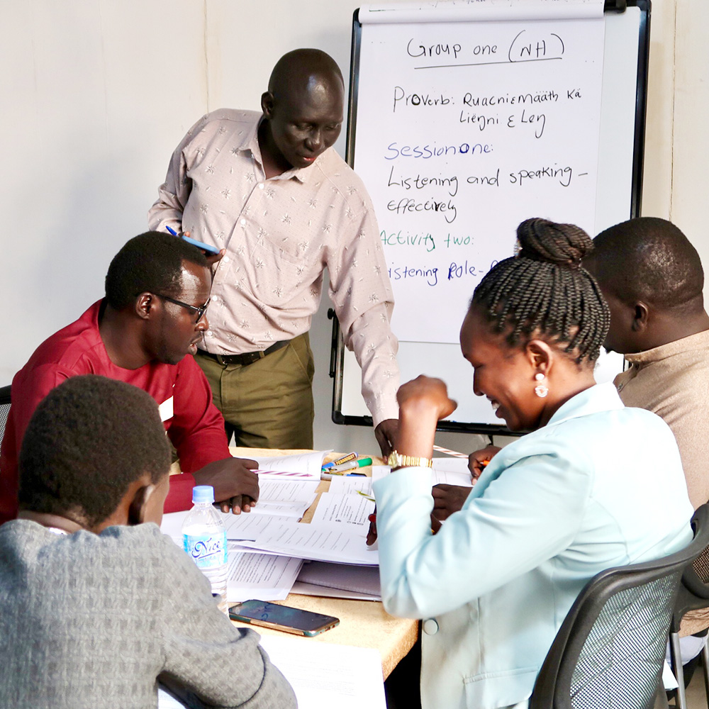 People seated at a table, with one standing and helping in front of a whiteboard.