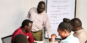 People seated at a table, with one standing and helping in front of a whiteboard.