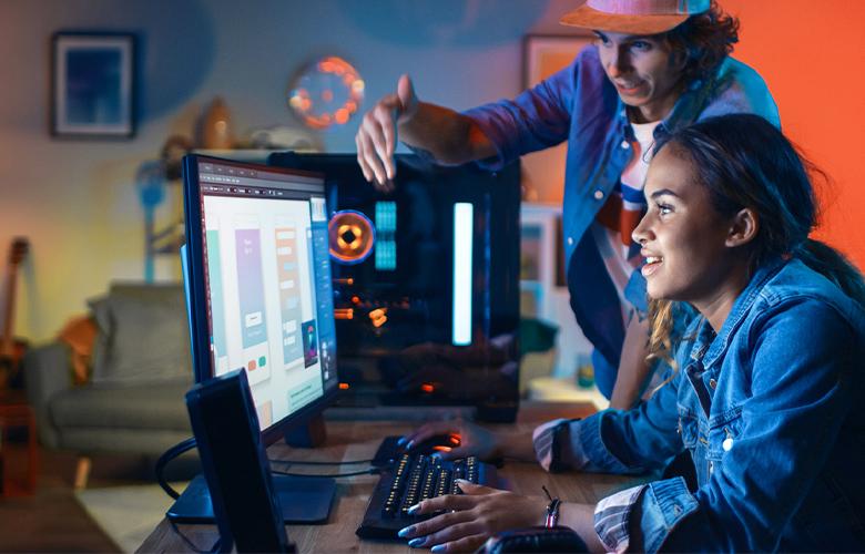 A younger woman working at a computer, with an adult standing beside her, in a technology learning environment.