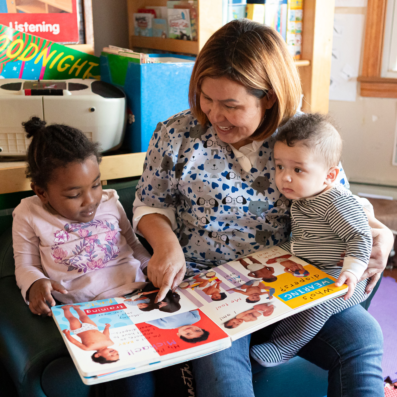 An older woman holding two small children on her lap, reading a book to them, in an educational setting.