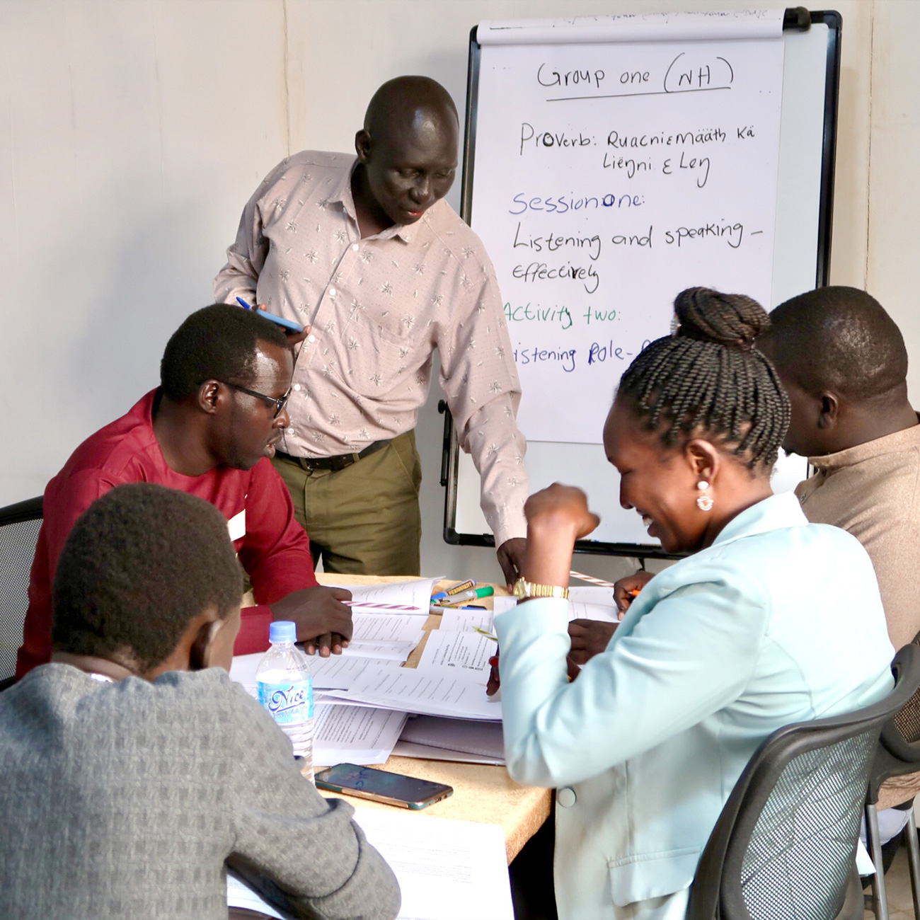 An educator stands with four individuals around a table, interacting with a learning easel pad in a collaborative setting.