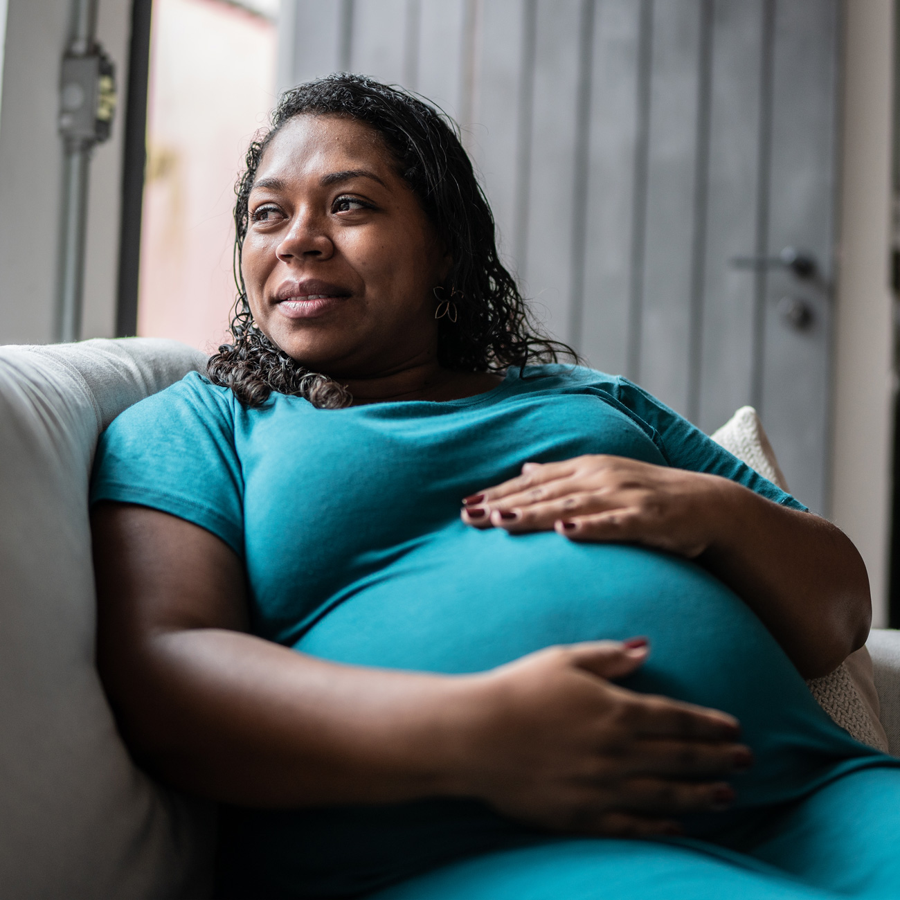 Woman sitting on a couch, smiling and holding her pregnant belly.
