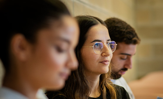 Three students seated in a row in a higher education classroom, with one looking outward and the other two looking down.