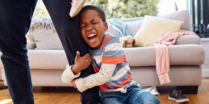 Young child grasping an adult's leg, mouth open as if crying, in a home setting.