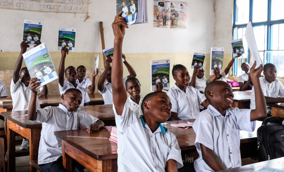 Youth with hands raised, wearing school uniforms, seated at desks in a primary-secondary classroom.